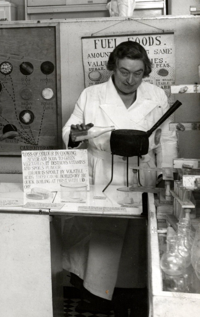 black and white photograph of woman pouring milk into a pan with a laboratory display around her.