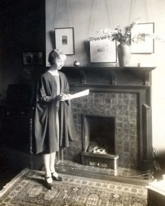 Black and white photograph of Dorothy Melvin standing at a fireplace reading a paper document