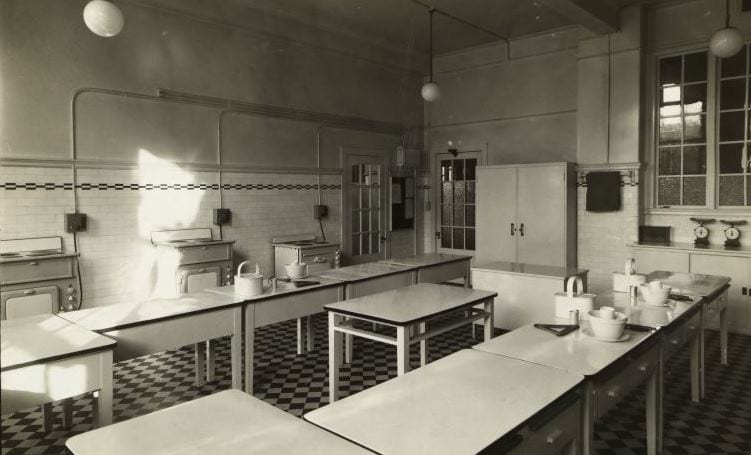 Black and white photograph of a teaching kitchen with 3 electric cookers along the back wall