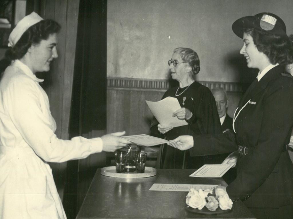 Black and white photograph of Princess Elizabeth handing a diploma to a student, with Dorothy Melvin in the background reading out names from a sheet.