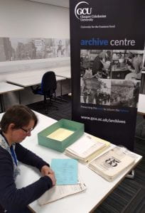 Colour photograph of Project Archivist working at a desk with bundles of papers with Archive Centre banner in the background.