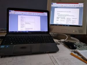 Photograph of a laptop and computer monitor on a table with papers and pencils beside it
