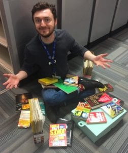 White man smiling at camera, seated. Books and magazines scattered around him